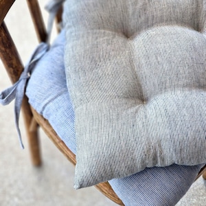 Two striped linen chair cushions stacked on a vintage wooden chair. The top cushion features a charcoal grey and white pinstripe pattern. The bottom cushion has a light blue and white stripe pattern. Both kitchen seat pads are tufted and include matching fabric ties.