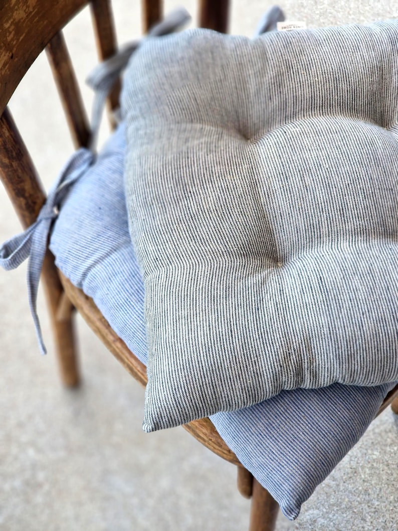 Two striped linen chair cushions stacked on a vintage wooden chair. The top cushion features a charcoal grey and white pinstripe pattern. The bottom cushion has a light blue and white stripe pattern. Both kitchen seat pads are tufted and include matching fabric ties.