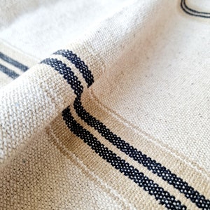 Pu&ograve; includere: Primo piano di un tessuto di lino beige con trama tessuta. Il tessuto presenta due strisce parallele blu navy e una striscia beige. Il tessuto &egrave; piegato, mostrando la trama e il motivo.