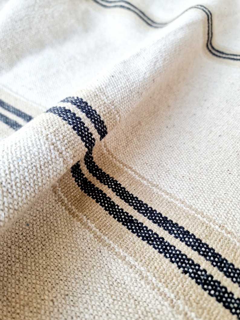 Pu&ograve; includere: Primo piano di un tessuto di lino beige con trama tessuta. Il tessuto presenta due strisce parallele blu navy e una striscia beige. Il tessuto &egrave; piegato, mostrando la trama e il motivo.