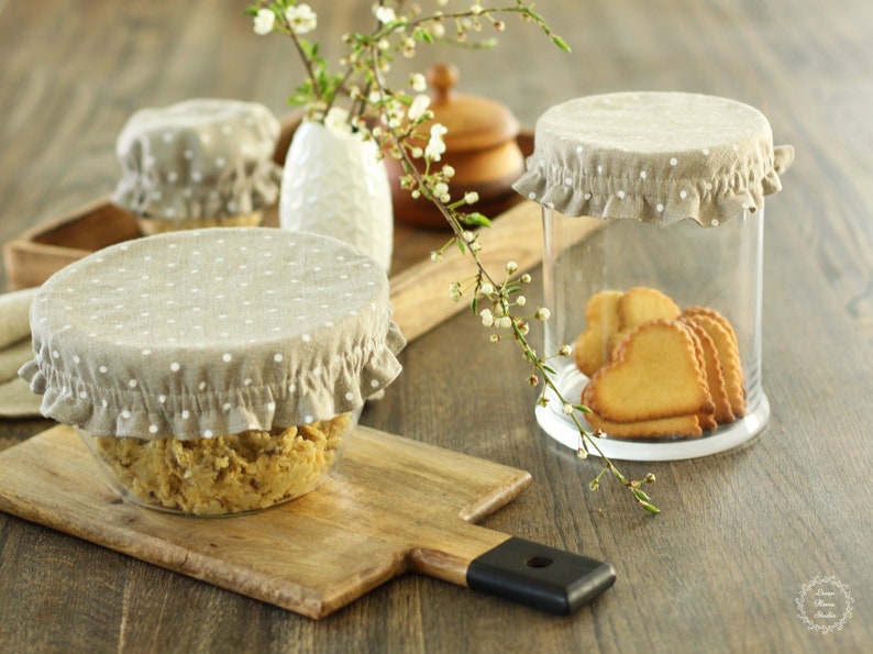 Reusable linen bowl and jar covers with a white polka dot pattern on natural fabric. Eco-friendly fabric lids with elastic ruffled edges on glass jars filled with cookies. Cozy rustic kitchen decor and a zero-waste alternative to plastic wrap.