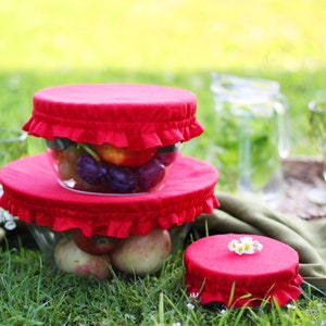 Bright red linen bowl covers on glass bowls filled with fresh fruit. Reusable fabric dish covers with elastic ruffles, displayed during an outdoor picnic on green grass. Eco-friendly summer kitchen accessories and sustainable food storage for outdoor dining.