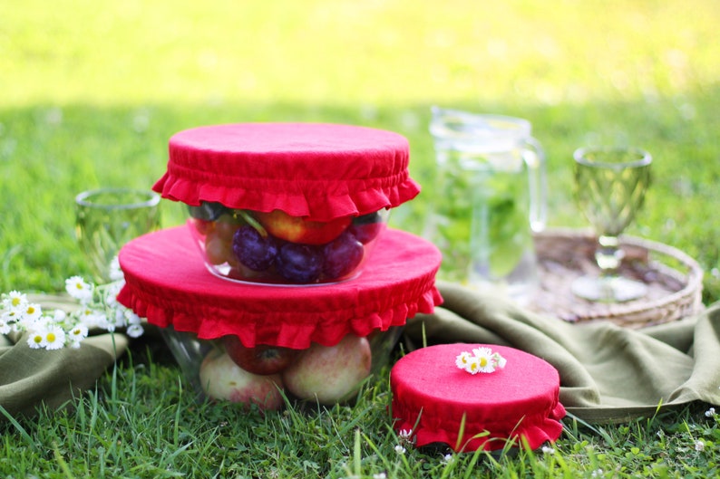 Bright red linen bowl covers on glass bowls filled with fresh fruit. Reusable fabric dish covers with elastic ruffles, displayed during an outdoor picnic on green grass. Eco-friendly summer kitchen accessories and sustainable food storage for outdoor dining.