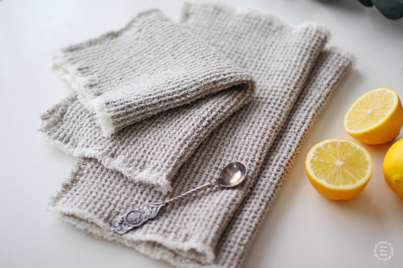 May include: Three waffle weave linen kitchen towels in a light gray color folded on a white surface with a silver spoon and two lemon halves.