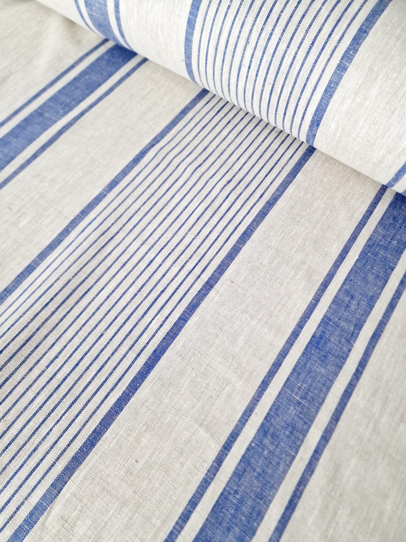Close-up of oatmeal-colored heavyweight linen fabric with vibrant royal blue grain sack stripes. The 100% natural flax linen is shown folded to highlight its thick, rustic texture and high-quality weave. Ideal for farmhouse style tablecloths, runners, or custom home decor