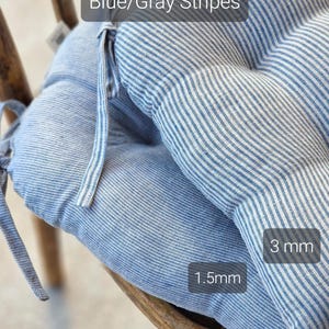 Close-up of two blue and gray striped linen chair cushions stacked on a wooden chair, showcasing different stripe widths. Text labels highlight the contrast: the bottom cushion has narrow 1.5mm stripes, and the top cushion has wider 3mm stripes. Both kitchen seat pads feature integrated fabric ties for securing to the chair.