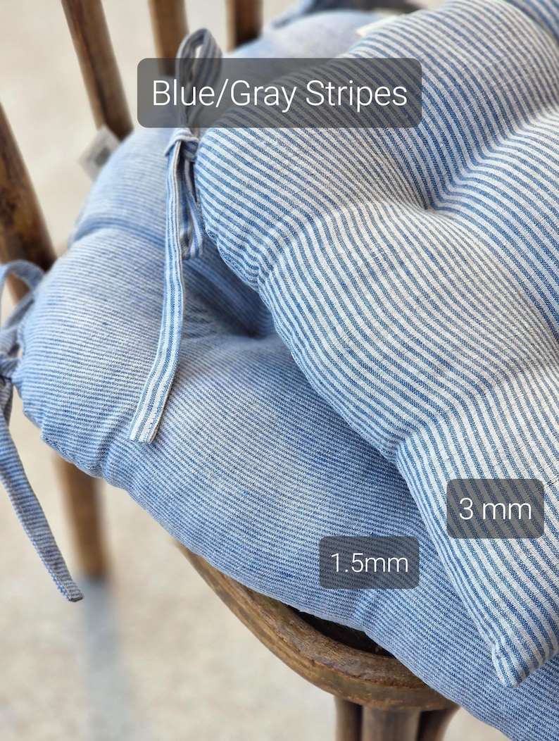 Close-up of two blue and gray striped linen chair cushions stacked on a wooden chair, showcasing different stripe widths. Text labels highlight the contrast: the bottom cushion has narrow 1.5mm stripes, and the top cushion has wider 3mm stripes. Both kitchen seat pads feature integrated fabric ties for securing to the chair.