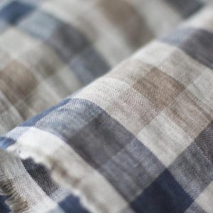 Close-up of a soft plaid linen fabric with a frayed edge, displaying a checkered pattern in navy blue, natural brown, beige, and white tones. The macro shot highlights the organic woven texture and slubs of the high-quality linen material.