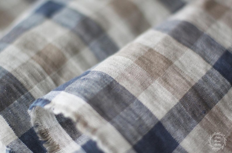 Close-up of a soft plaid linen fabric with a frayed edge, displaying a checkered pattern in navy blue, natural brown, beige, and white tones. The macro shot highlights the organic woven texture and slubs of the high-quality linen material.