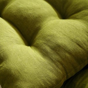 Close-up of a moss green linen chair cushion, showcasing the rich woven texture and natural slubs of the fabric. The image focuses on the plump, tufted sections and soft stitched dimples of the olive green seat pad.