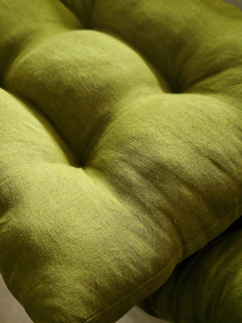 Close-up of a moss green linen chair cushion, showcasing the rich woven texture and natural slubs of the fabric. The image focuses on the plump, tufted sections and soft stitched dimples of the olive green seat pad.