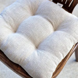 A square, tufted linen chair cushion in a natural oatmeal or flax color, placed on a dark wooden dining chair. The rustic farmhouse style kitchen seat pad shows a rich woven fabric texture and includes integrated ties at the back.