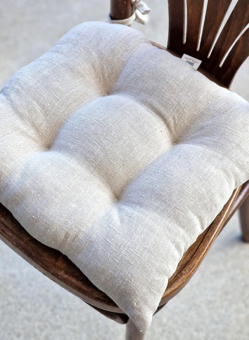 A square, tufted linen chair cushion in a natural oatmeal or flax color, placed on a dark wooden dining chair. The rustic farmhouse style kitchen seat pad shows a rich woven fabric texture and includes integrated ties at the back.