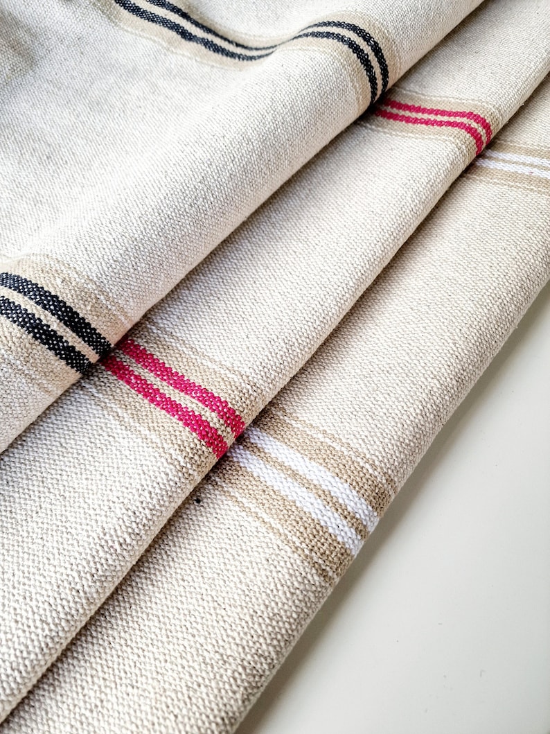 Pu&ograve; includere: Primo piano di tessuto di lino piegato con accenti a righe. Il tessuto &egrave; di colore beige naturale, con strisce nere, rosa e bianche. Il tessuto ha un aspetto strutturato, che suggerisce una costruzione tessuta. Le strisce sono uniformemente distanziate.