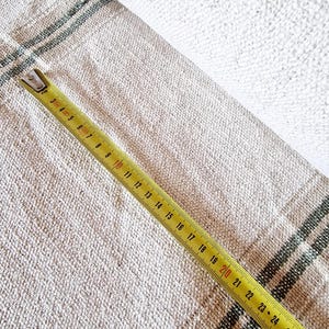 Pu&ograve; includere: Tessuto di lino beige con strisce verde scuro parzialmente arrotolato. Un metro a nastro giallo, che mostra le misure in centimetri, &egrave; appoggiato sul tessuto. Il tessuto ha un aspetto strutturato.
