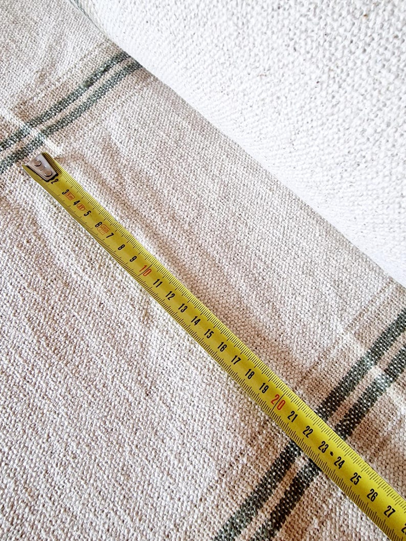 Pu&ograve; includere: Tessuto di lino beige con strisce verde scuro parzialmente arrotolato. Un metro a nastro giallo, che mostra le misure in centimetri, &egrave; appoggiato sul tessuto. Il tessuto ha un aspetto strutturato.