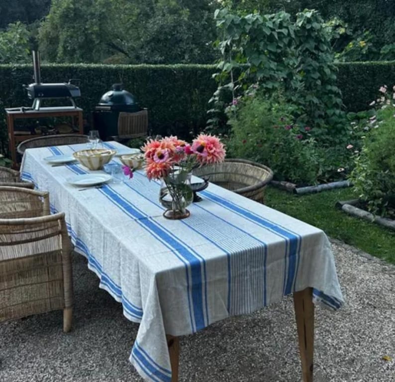 Full view of a long, rectangular royal blue striped linen tablecloth on an outdoor patio table. The table is set with pink dahlias in a vase and ceramic bowls, surrounded by wicker chairs in a lush green garden setting. Perfect for summer garden parties and rustic outdoor dining.