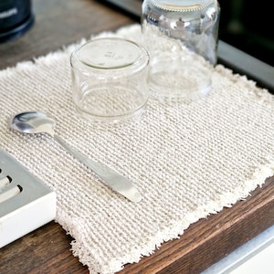 May include: A white, textured kitchen towel with two clear glass jars and a silver spoon on top. The towel is on a wooden surface.