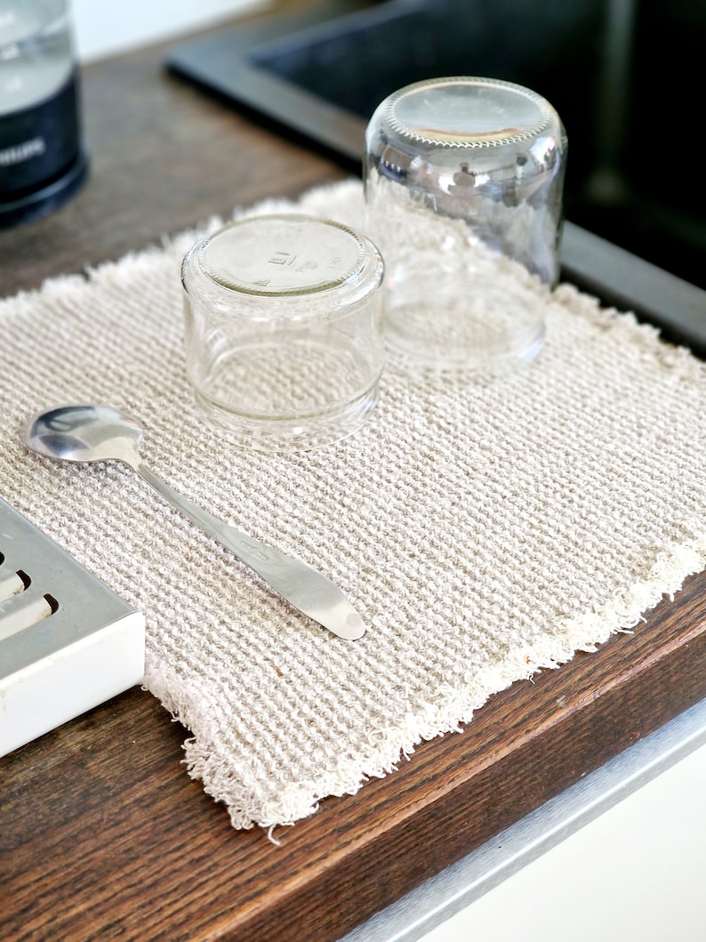 May include: A white, textured kitchen towel with two clear glass jars and a silver spoon on top. The towel is on a wooden surface.