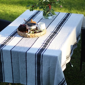 Striped linen tablecloth on an outdoor garden table set on green grass. Light grey natural linen fabric with navy blue grain sack stripes. A lifestyle scene featuring a wildflower bouquet in a glass vase and a wicker tray with a teapot in soft afternoon sunlight