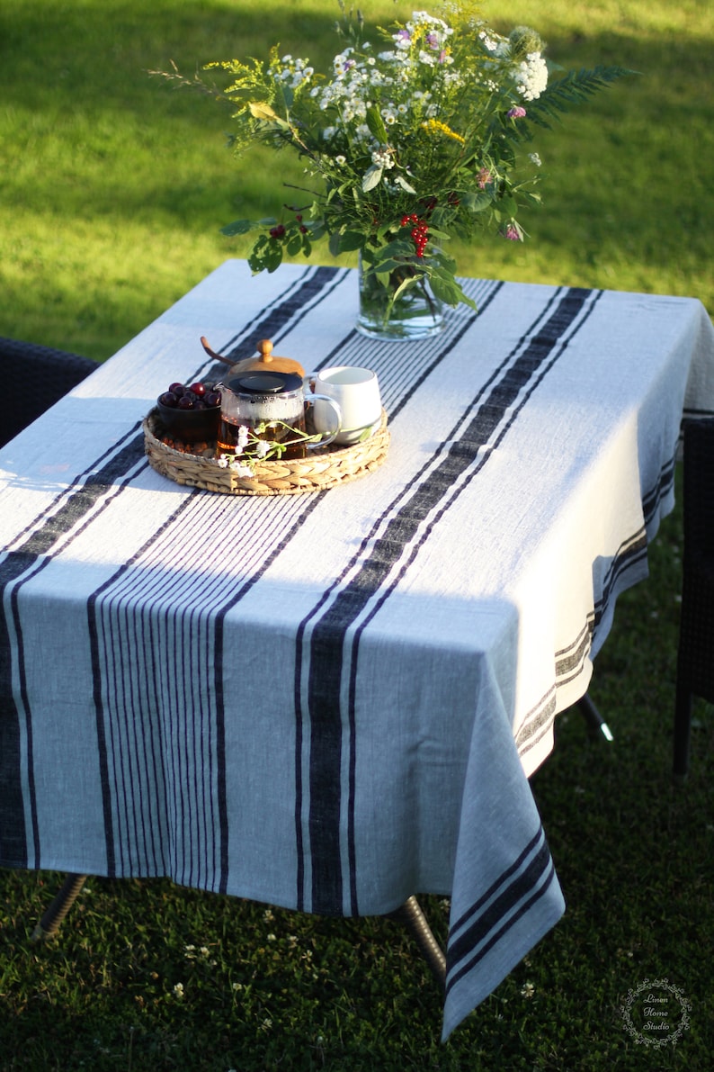 Striped linen tablecloth on an outdoor garden table set on green grass. Light grey natural linen fabric with navy blue grain sack stripes. A lifestyle scene featuring a wildflower bouquet in a glass vase and a wicker tray with a teapot in soft afternoon sunlight
