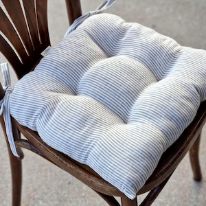 A square, tufted linen chair cushion with a grey and white striped pattern, placed on a dark wooden dining chair. The farmhouse style kitchen seat pad is plumply filled and features matching fabric ties secured to the chair back.