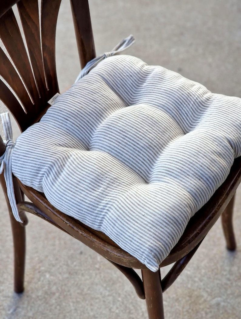 A square, tufted linen chair cushion with a grey and white striped pattern, placed on a dark wooden dining chair. The farmhouse style kitchen seat pad is plumply filled and features matching fabric ties secured to the chair back.