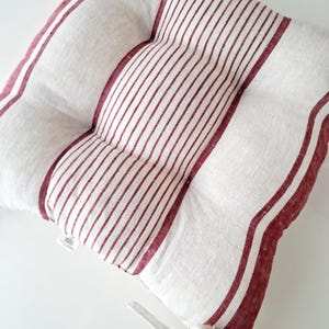 May include: A square cushion with a white background and a red striped pattern. The cushion has a tufted design and a white tie on one side. The stripes are vertical in the center and horizontal on the edges.