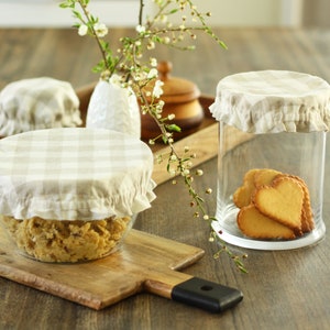 Natural beige and white gingham checkered linen bowl covers. Set of reusable fabric jar lids with elastic ruffled edges on glass containers, styled with heart-shaped cookies and spring blossoms. Classic farmhouse kitchen decor and zero-waste gift idea.
