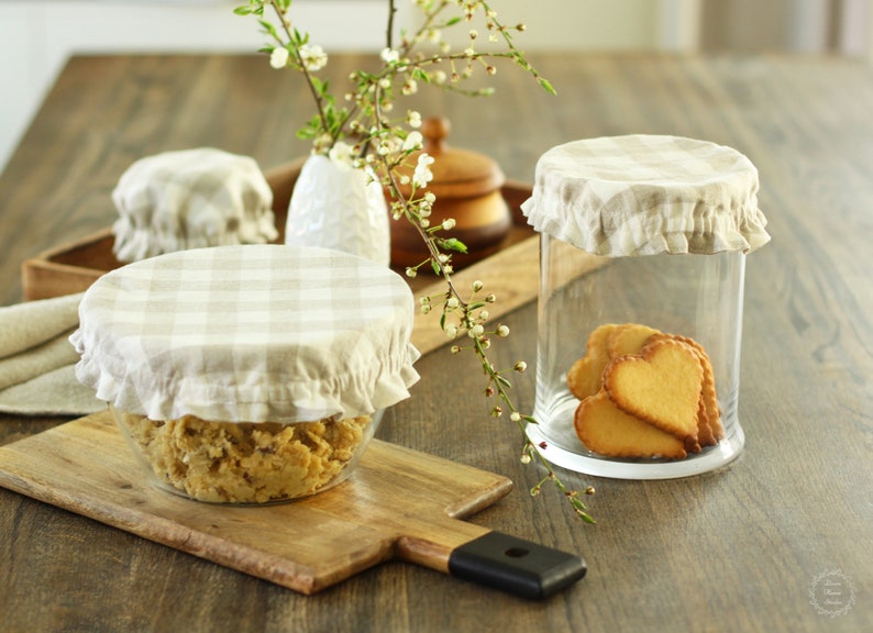 Natural beige and white gingham checkered linen bowl covers. Set of reusable fabric jar lids with elastic ruffled edges on glass containers, styled with heart-shaped cookies and spring blossoms. Classic farmhouse kitchen decor and zero-waste gift idea.