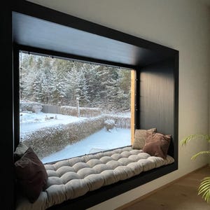 May include: A cozy window seat with a tufted cushion and two throw pillows. The window frame is black, and the view outside shows a snowy landscape with evergreen trees. A potted plant sits nearby.