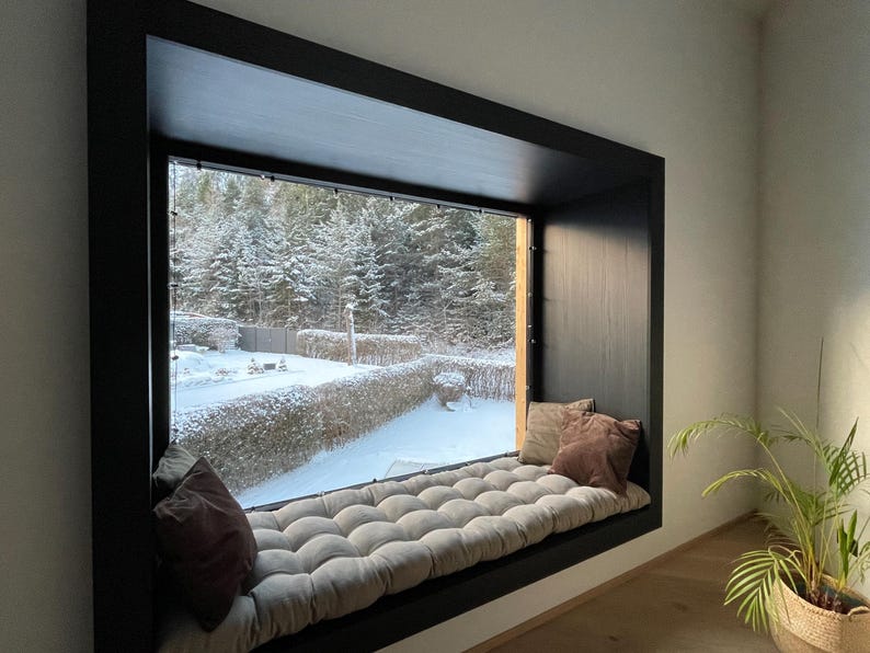 May include: A cozy window seat with a tufted cushion and two throw pillows. The window frame is black, and the view outside shows a snowy landscape with evergreen trees. A potted plant sits nearby.