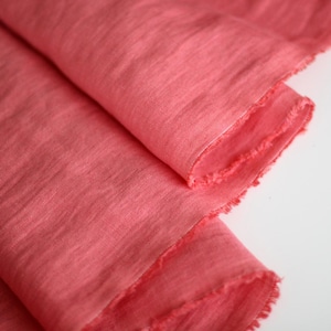 May include: Close-up of coral-colored fabric with a raw, frayed edge. The fabric is folded over, showing the texture and color variations. The material appears soft and lightweight, suitable for apparel or craft projects.