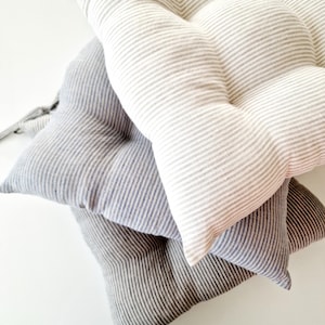 A stack of three striped linen chair cushions in different neutral shades on a white surface. The top cushion has a light beige and white stripe pattern, the middle one is light blue and white striped, and the bottom one is dark grey and white striped. All kitchen seat pads are tufted and plump.