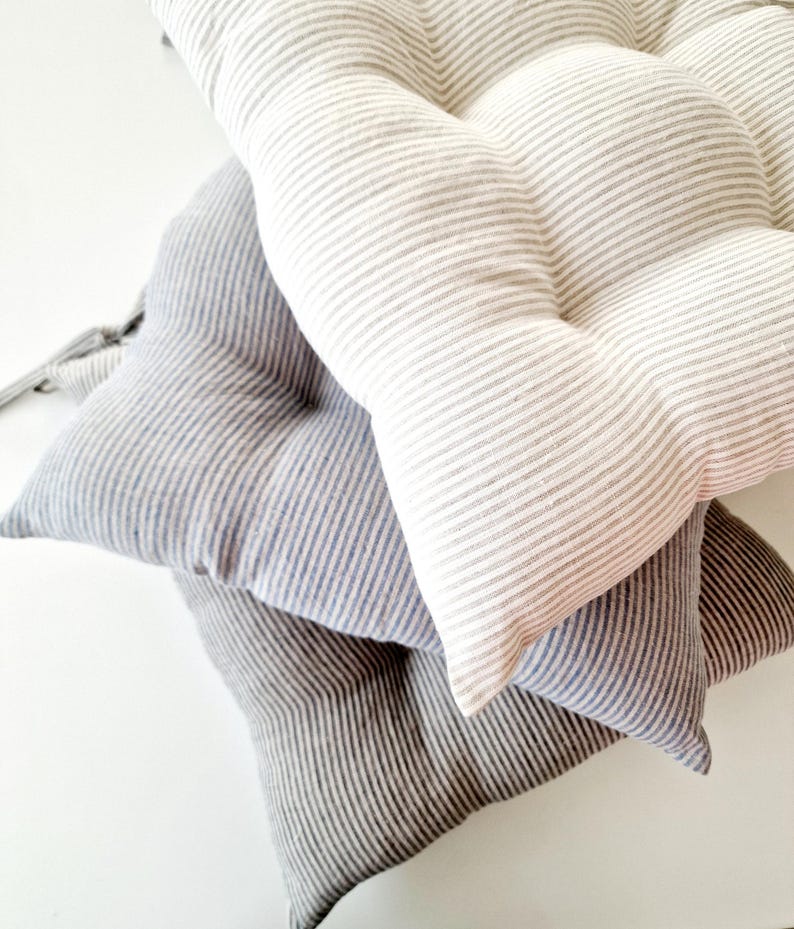 A stack of three striped linen chair cushions in different neutral shades on a white surface. The top cushion has a light beige and white stripe pattern, the middle one is light blue and white striped, and the bottom one is dark grey and white striped. All kitchen seat pads are tufted and plump.