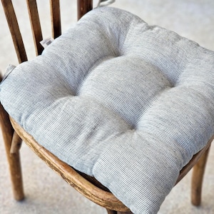 A square, tufted linen chair cushion with a charcoal grey and off-white striped pattern, placed on a vintage wooden dining chair. The farmhouse style kitchen seat pad features integrated fabric ties attached to the back of the chair.
