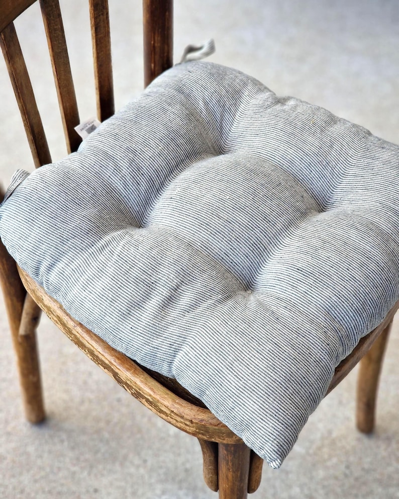 A square, tufted linen chair cushion with a charcoal grey and off-white striped pattern, placed on a vintage wooden dining chair. The farmhouse style kitchen seat pad features integrated fabric ties attached to the back of the chair.