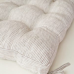 May include: A tufted cushion with a striped pattern in shades of white and gray. The cushion has a rounded shape and is tied with a matching fabric ribbon. The fabric has a soft, textured appearance, suitable for seating or decorative use.