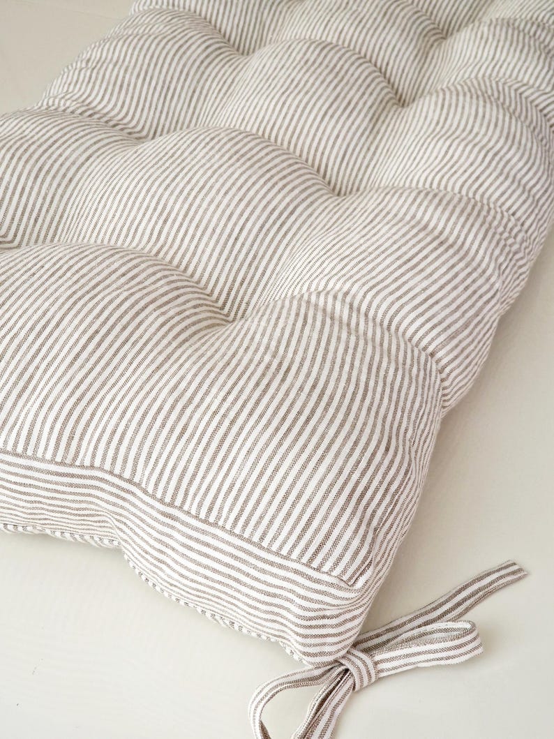 May include: A tufted cushion with a striped pattern in shades of white and gray. The cushion has a rounded shape and is tied with a matching fabric ribbon. The fabric has a soft, textured appearance, suitable for seating or decorative use.