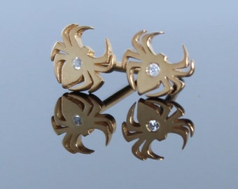 May include: A pair of gold-colored earrings, each shaped like a stylized spider with curved legs. A small, clear gemstone is set in the center of each spider. The earrings are displayed on a reflective surface.