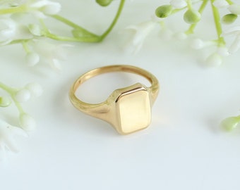 May include: A gold signet ring with a rectangular face. The ring is made of polished gold and has a smooth, rounded band. The ring is shown on a white surface with white flowers in the background.