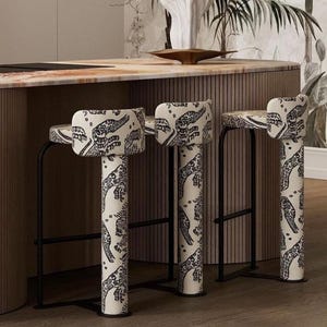 May include: Three modern bar stools with black metal frames and patterned fabric seats and legs. The fabric features a black and white design. The stools are positioned in front of a wooden kitchen island with a marble countertop.