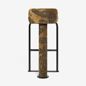 May include: A tall, upholstered stool with a rectangular seat and a cylindrical support. The seat and support are covered in a mustard-colored fabric with a dark gray pattern. The stool has a black metal frame.