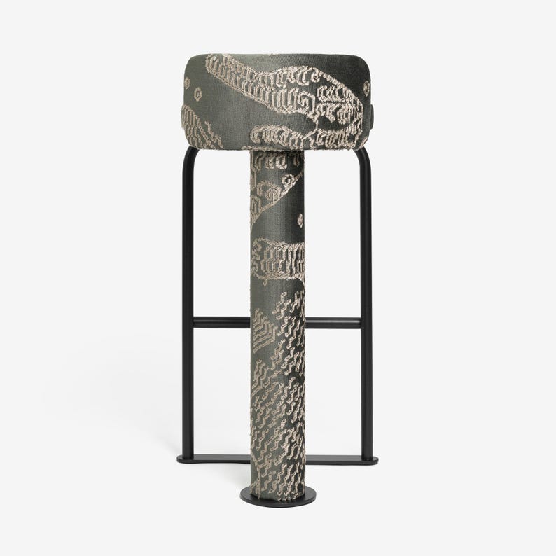 May include: A modern bar stool with a rectangular, upholstered seat and a patterned design in shades of gray and beige. The stool has a black metal frame and a cylindrical central support. The overall design is contemporary and stylish.