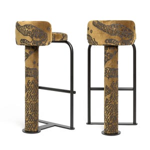 May include: Two bar stools with black metal frames and upholstered seats and columns. The upholstery is a mustard yellow fabric with a dark gray pattern. The stools have a minimalist design, suitable for a kitchen or bar area.