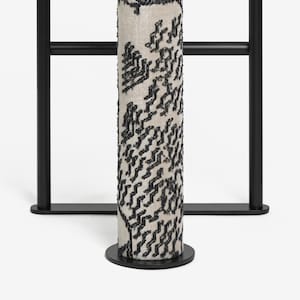 May include: A cylindrical, cream-colored fabric column with a black, textured pattern. The column is mounted on a black circular base and set against a black metal frame. The design is modern and minimalist.
