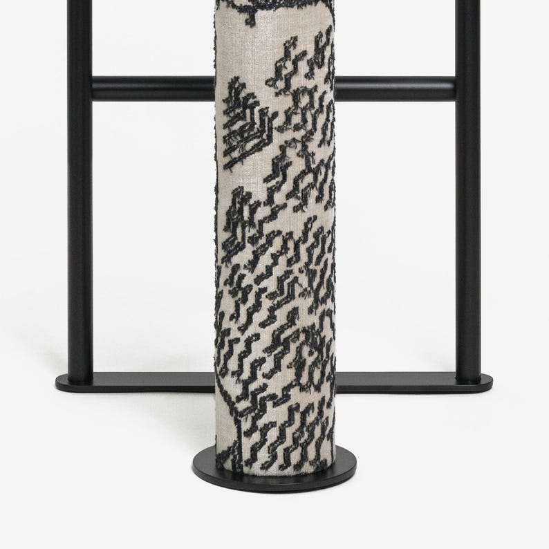 May include: A cylindrical, cream-colored fabric column with a black, textured pattern. The column is mounted on a black circular base and set against a black metal frame. The design is modern and minimalist.