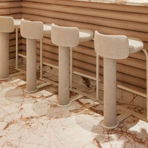May include: A row of four beige upholstered bar stools with rounded backs and cylindrical legs. The stools are set against a wooden bar with a marble countertop. The floor is marble with brown and beige veining.