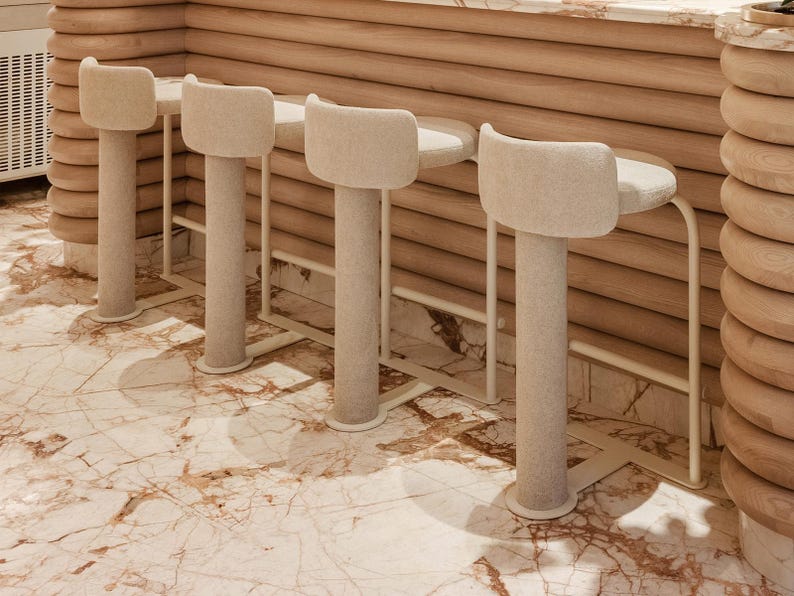 May include: A row of four beige upholstered bar stools with rounded backs and cylindrical legs. The stools are set against a wooden bar with a marble countertop. The floor is marble with brown and beige veining.