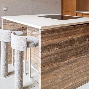 May include: A modern kitchen island with a white countertop and a built-in cooktop. The island base is made of brown, textured stone. Two white, upholstered bar stools with curved backs and white metal legs are positioned in front of the island.